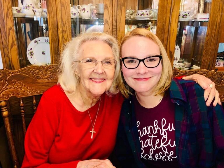 My grandma wearing a red sweater sitting at a table embracing me as I wear a burgundy shirt with a green flannel top over it.