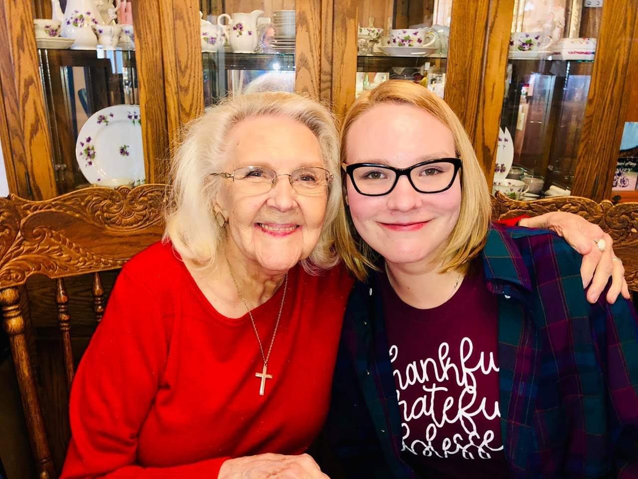 My grandma wearing a red sweater sitting at a table embracing me as I wear a burgundy shirt with a green flannel top over it.